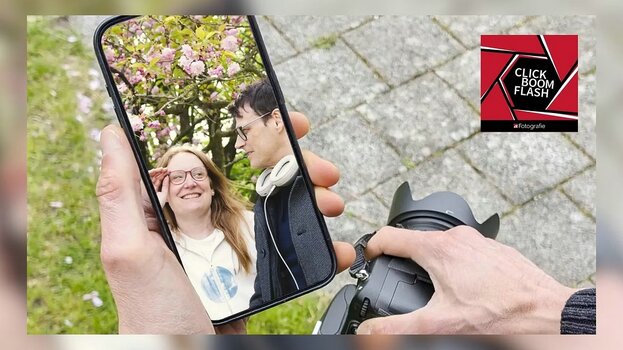 Eine Person hält ein Smartphone und fotografiert ein Paar unter einem blühenden Baum; im Vordergrund hält eine andere Person eine Spiegelreflexkamera – ein Symbol für den Vergleich zwischen Smartphone und klassischer Fotografie.