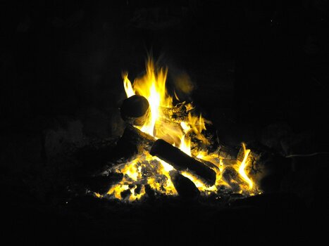 Ein kleines Lagerfeuer bei Nacht mit brennenden Holzscheiten, hellen gelben Flammen und glühender Asche in der Dunkelheit.