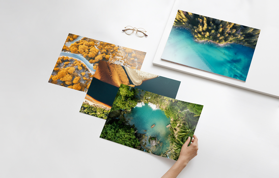 Nahaufnahme einer Hand, die einen Foto-Print einer tropischen Lagune hält, im Hintergrund ein Stapel weiterer hochwertiger Naturaufnahmen und ein gerahmtes Wandbild.
