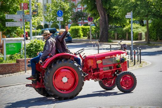 Zwei Männer mit Sonnenhüten sitzen auf einem roten Oldtimer-Traktor, der mit weißen Blumen geschmückt ist und über eine sonnige Straße fährt.