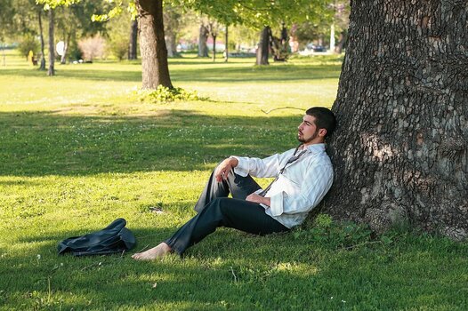 Ein entspannt wirkender junger Mann sitzt barfuß im Gras und lehnt an einem großen Baumstamm im kühlen Schatten, während im Hintergrund eine sonnendurchflutete Parklandschaft zu sehen ist.
