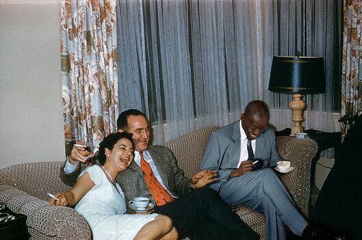 Inszenierte Farbfotografie im Stil privater Aufnahmen der 1950er-Jahre: Eine Frau im weißen Kleid lacht ausgelassen auf einem Sofa, während zwei Männer im Anzug neben ihr Tee oder Kaffee trinken und rauchen.
