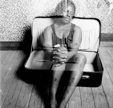 Eine melancholische Schwarz-Weiß-Fotografie im Doppelbelichtungs-Stil, die eine transparente Frauenfigur sitzend in einem großen, geöffneten Überseekoffer auf Holzdielen zeigt. Eine melancholische Schwarz-Weiß-Fotografie im Doppelbelichtungs-Stil, die eine transparente Frauenfigur sitzend in einem großen, geöffneten Überseekoffer auf Holzdielen zeigt.