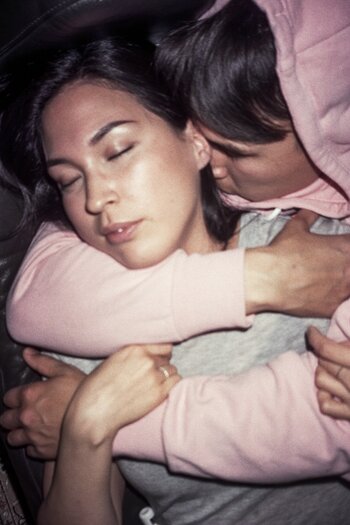 Ein nahes, farbiges Porträt im sanften Licht, das zwei eng umschlungene Menschen zeigt, wobei einer eine rosa Kapuze trägt und den Kopf an den Hals des anderen schmiegt. Ein nahes, farbiges Porträt im sanften Licht, das zwei eng umschlungene Menschen zeigt, wobei einer eine rosa Kapuze trägt und den Kopf an den Hals des anderen schmiegt.