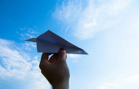 Hand hält einen Papierflieger vor blauem Himmel mit leichten Wolken