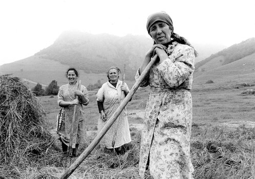 Eine Schwarz-Weiß-Reportageaufnahme von Franki Raffles aus der Sowjetunion (1989): Drei Frauen in ländlicher Arbeitskleidung und mit schützenden Kopftüchern stehen mit Heugabeln in den Händen auf einem Feld. Im Hintergrund ist eine weite, leicht hügelige Landschaft zu sehen.