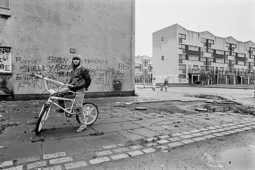 ): Schwarz-Weiß-Fotografie von Franki Raffles: Ein Mädchen mit Fahrrad steht vor einer graffitibedeckten Häuserwand in einer trostlosen Wohnsiedlung, während im Hintergrund weitere Wohnblöcke und Passanten zu sehen sind.