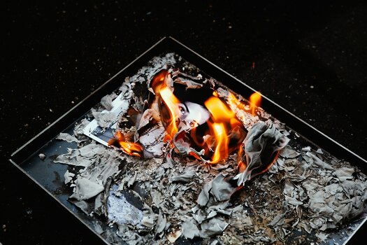 Eine dunkle Metallwanne voller Asche und verbrannter Fotografien, aus denen helle orangefarbene Flammen aufsteigen. Zwischen den verkohlten Bildresten ist noch das Gesicht einer porträtierten Frau erkennbar.
