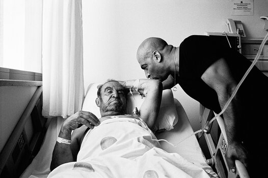 Intime Schwarz-Weiß-Fotografie aus „Rotting from Within“: Eine Person verabschiedet sich am Krankenbett eines älteren Familienmitglieds. Intime Schwarz-Weiß-Fotografie aus „Rotting from Within“: Eine Person verabschiedet sich am Krankenbett eines älteren Familienmitglieds.