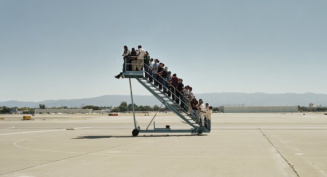Mehrere Menschen stehen dicht gedrängt auf einer Flugzeugtreppe, die ins Leere führt, in Adrian Pacis Arbeit „Centro di permanenza temporanea“.