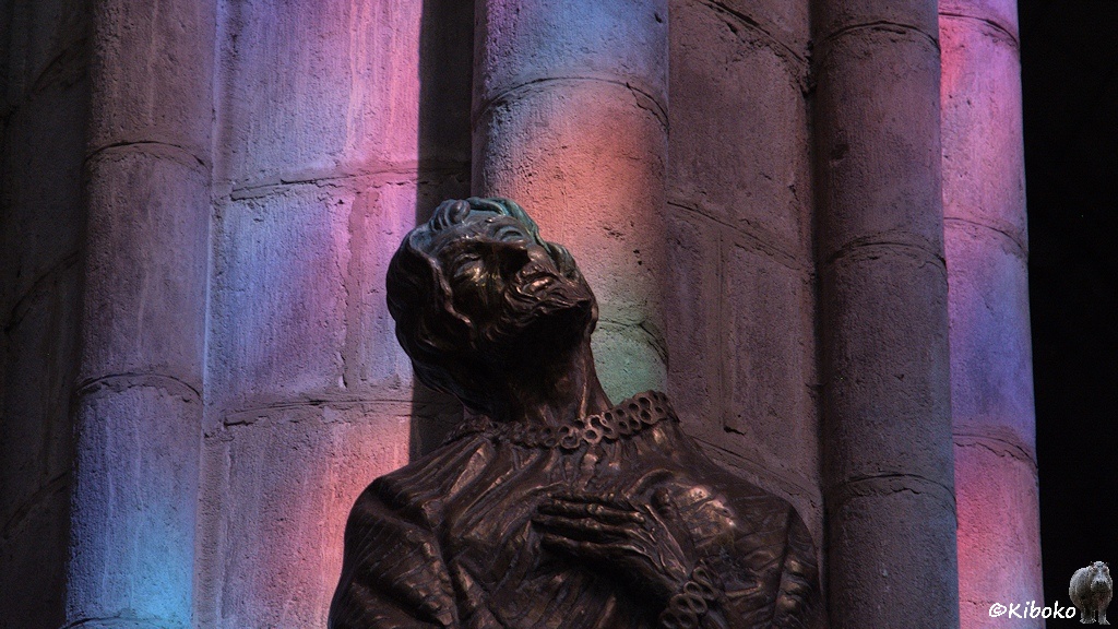 Das Bild zeigt das Porträt einer Figure vor einer Säule. Die Sonne scheint durch ein buntes Glasfenster und beleuchtet die Säulöe in rosa, blau, lila, orang eund grün.