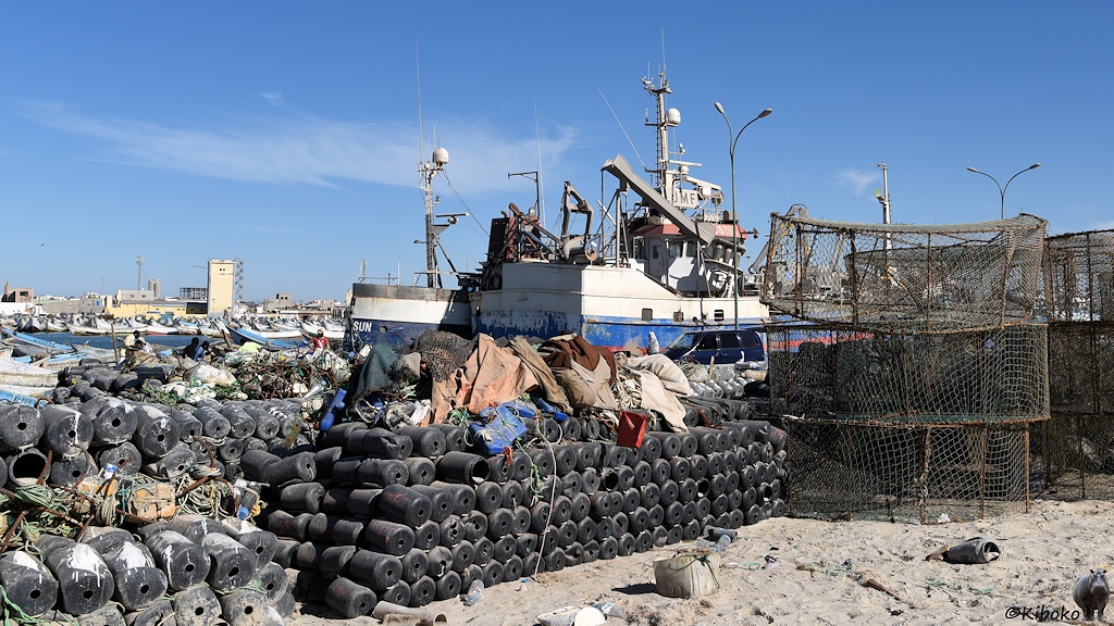 Das Bild zeigt schwarze, runde Plastikschwimmer, die in langen Reihen sechsfach gestapelt sind. Daneben stehen Drahtkäfige, die dreifach gestapelt sind. Im Hintergrund sind weiß-blau-rot lackierte Fischtrawler.