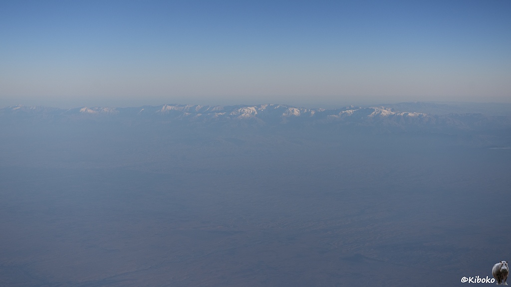Das Bild zeigt eine Luftaufnahme auf einer Gebirgskette mit schneebedeckten Gipfeln. Die Berge und die Ebene davor liegen in einen bläulichen Dunst.
