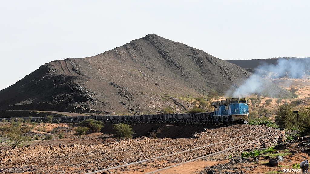 Zwei hellblaue Dieselloks scheben die leeren Erzwagen an einem dunkelgrauen Berg vorbei und stoßen dabei hellblaue Abgaswolken aus.