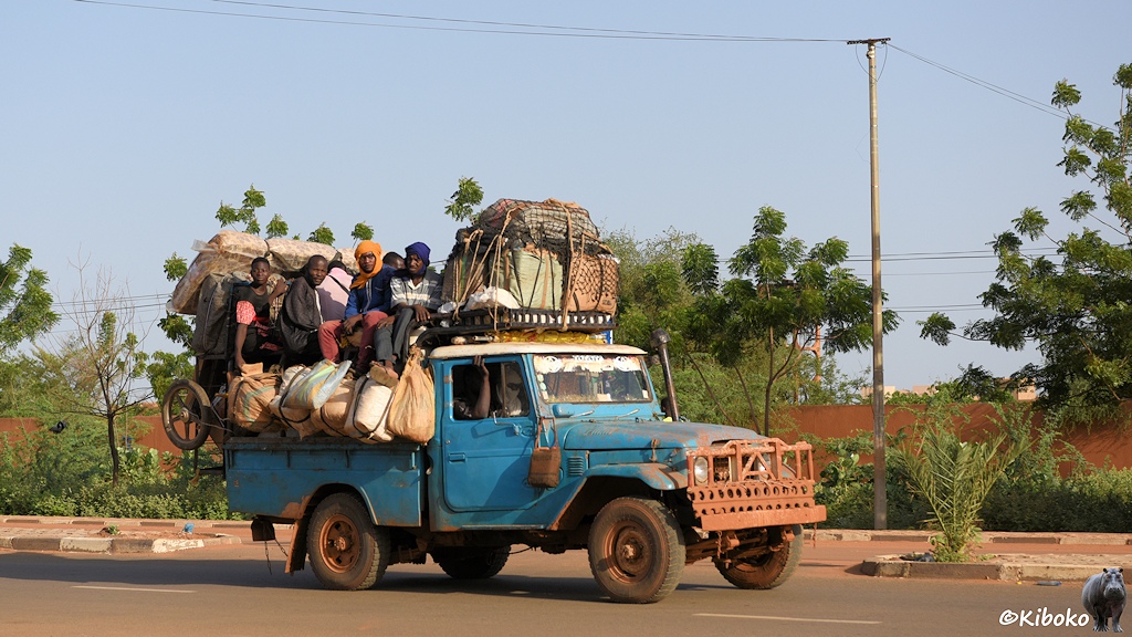 Das Bild zeigt einen alten, himmelblauen Toyota Geländewagen mit Pritsche. Vorn iat ein massiver Rammschutz montiert. Auf dem Kabinendach ist ein großer Haufen Koffer. Dahinter hocken ca. 10 Männer auf der Ladung. An der Seite sind angebundene Beutel und Säcke. Hinten sind mehrere Matratzen angebunden.