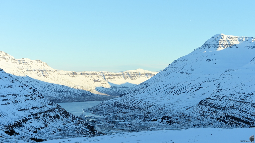 Blick von einem Berg auf ein Fjordende, das von schneebedeckten Bergen umgeben ist. Nur die oberen Teile der Berghänge sind auf einer Seite in der Sonne. Das Tal liegt im Schatten.