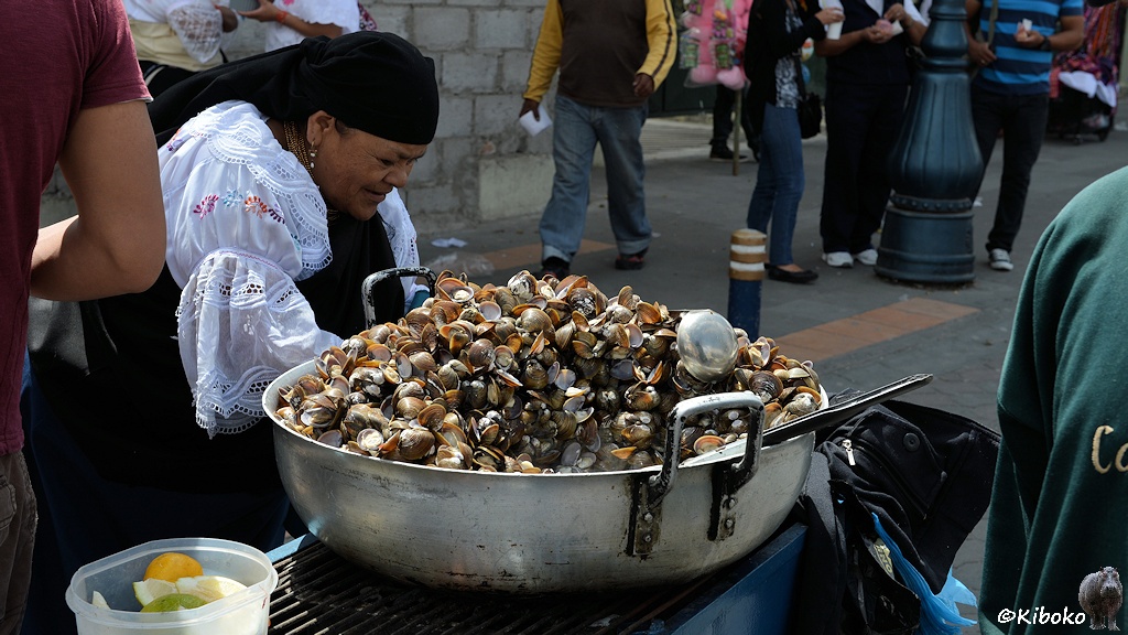 Das Bild zeigt eine ältere Dame mit weißgestickter Bluse, schwarzem Kopftuch und schwarzem Rock in gebückter Haltung vor einem Grill. Auf dem Grill steht eine sehr große silberne Schüssel. Die ist übervoll mit einen Berg aus offenstehenden Muschlen. Eine silberne Kelle liegt auf dem Berg. Daneben stehen in einer Plastikschüssel Zitronenscheiben.