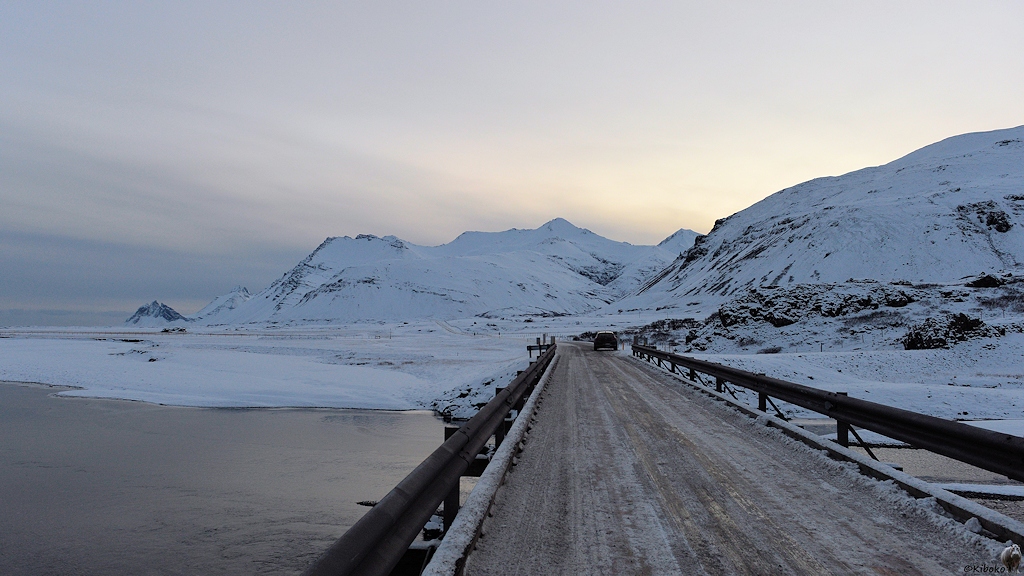 Das Bild zeigt eine einspurige Brücke über einen Fluss. Im Hintergrund sind schneebedeckte Berge.
