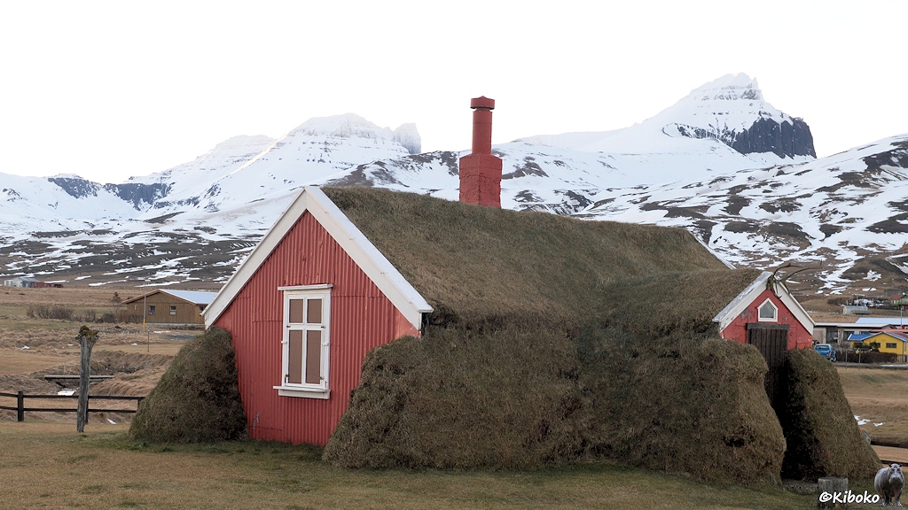 Auf einer Wiese steht ein kleines Haus mit Grasdach und grasbewachsenen Wänden. Die Giebel sind aus rotbraun gestrichenen Holz. Die Fenster und die Giebelkante sind weiß lackiert. Aus dem Dach ragt ein rotbrauner Schornstein. Im Hintergrund sind schneebedeckte Berge.