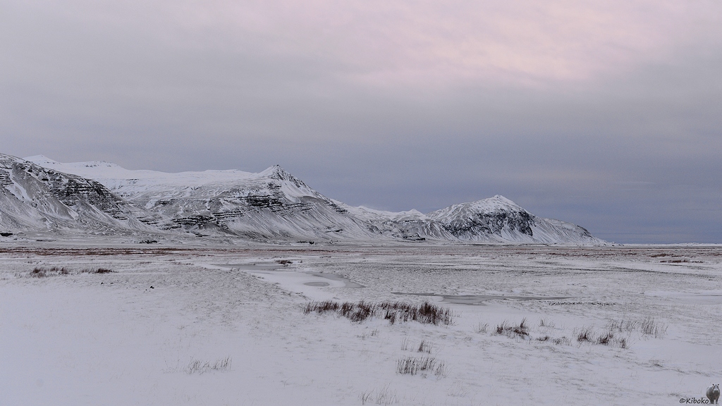 Das Bild zeigt eine verschneite Schwemmsandebene. Dahinter ist eine Bergkette.