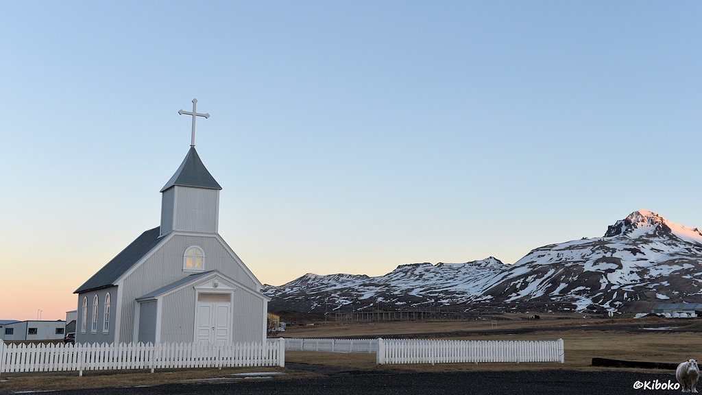 Eine kleine hellgraue Holzkirche mit drei Seitenfenstern und Miniturm steht auf einer Wiese. Die Wiese ist von einem weißen Lattenzaun eingefasst. Im Hintergrund sind schneebedeckte Berge.