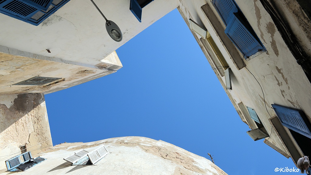 Das Bild zeigt den Blick aus einer engen Gasse nach oben. Die weißen Häuser mit bleuan Fensterrahmen rahemn den Blick zum Himmel ein. Die Form ist ein Dreieck, dass mit der Spitze nach oben zeigt. Die untere Kannte iste in Rundbogen.