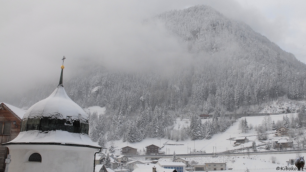 Das Bild zeigt eine Landschaftsaufnahme von einem kleinen Zug im Gebirge. Im Vordergrund steht links ein weißer Turm einer Kriche mit zwiebelförmigen Kupferdach. Dahinter ist ein verschneiter Berghang mit Nadelbäumen.