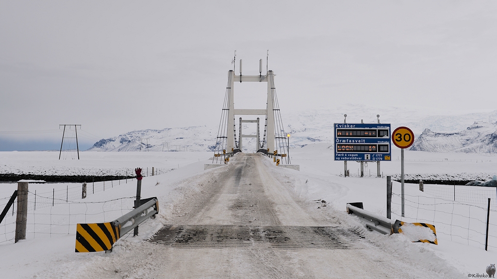 Das Bild zeigt die verschneite Straße über eine Hängebrücke mit zwei weißen Pylonen. Davor ist ein Viehgatter in der Straße eingelassen. Ein blaues Hinweisschild mit Leuchtanzeige informiert über Temperatur und Windgeschwindigkeiten. Ein 30 km/h Schild mit gelbem Hintergrund steht ebenfalls am Straßenrand.
