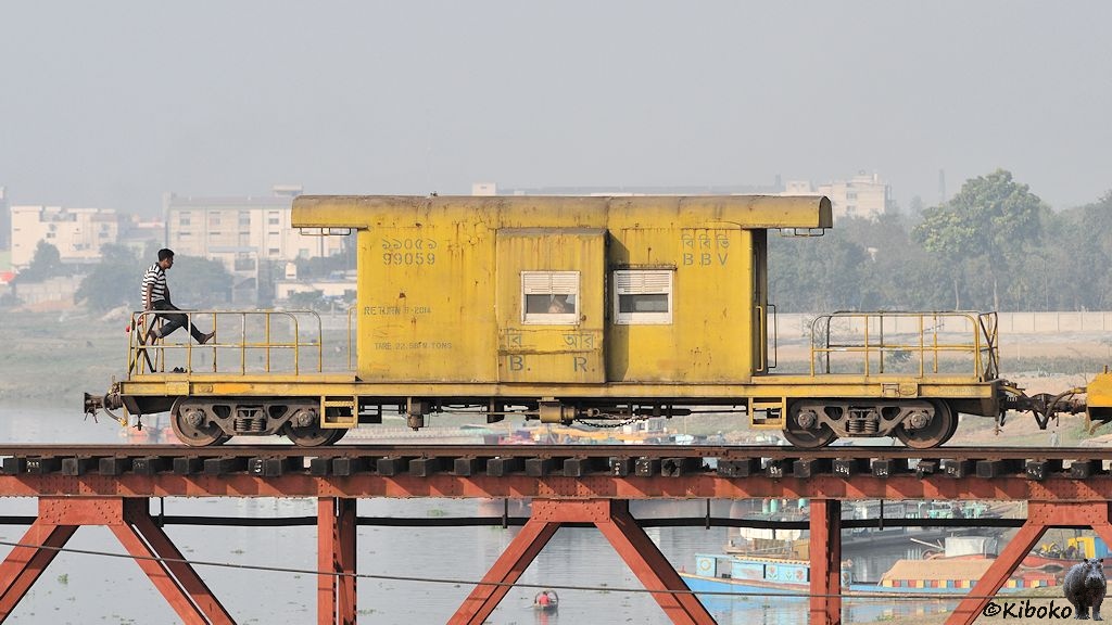 Das Bild zeigt einen gelben vierachsigen Güterzugbegleitwagen am Ende eines Zuges auf einer Untergurtträgerbrücke. Es ist ein flacher Wagen mit einer Blechhütte in der Mitte. Die Enden sind mit Geländer gesichert. Auf dem Geländer sitzt ein Mitfahrer und lässt die Beine baumeln.