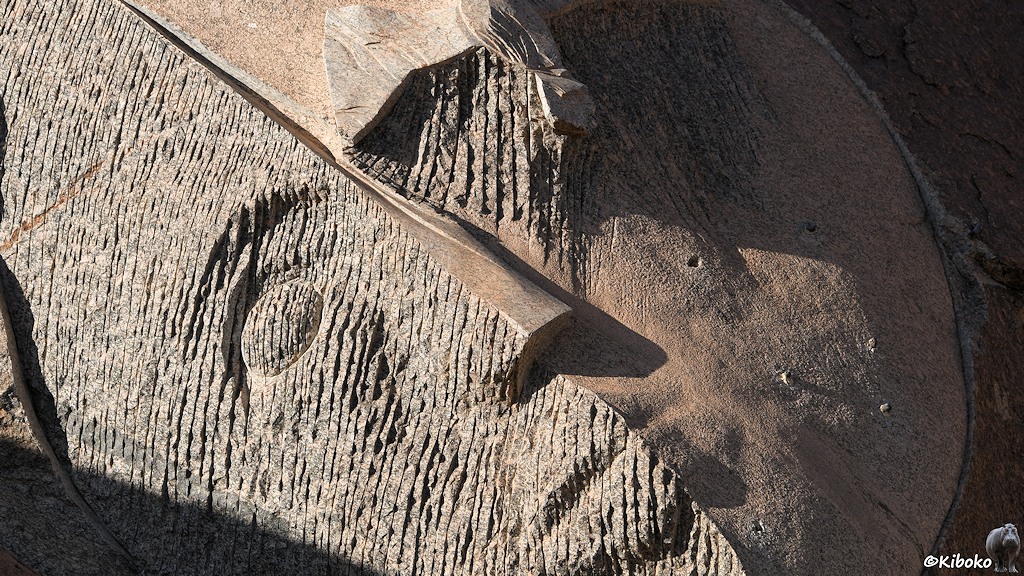 Das Bild zeigt ein in den Felsen gemeißeltes Gesicht. Das Gesicht ist diagonal nach links geneigt. Die linke Hälfte hat vertikal Riefen, während die rechte Hälfte glatt dargestellt ist.