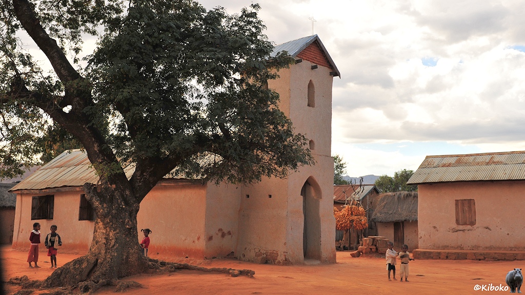 Das Bild zeigt eine kleine weiße Kirche mit viereckigen Turm über dem Eingang. Die Kirche steht auf einen Platz, der mit rötlichem Sand bedeckt ist. Vor und neben der Kirche spielen 5 kleine Kinder. Neben der Kriche steht ein großer Baum mit dicken, kurzen Stamm und ausladenden Ästen.