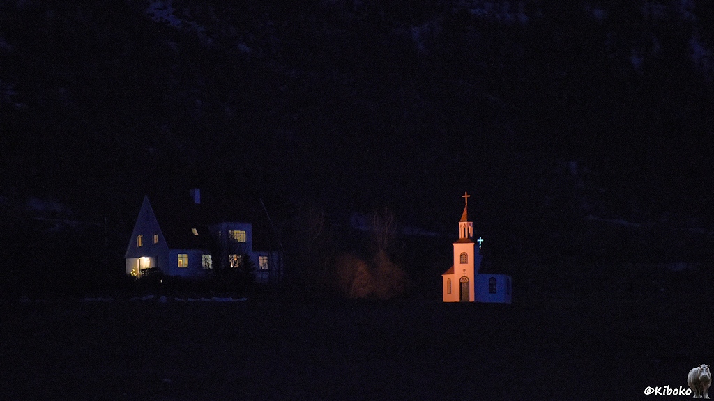 An einem Berghang stehen in der Dunkelheit ein Haus und eine Kirche. Das Haus ist größer als die Kirche. Beim Haus sind alle Fenster beleuchtet. Die Kirche ist von außen angestrahlt.
