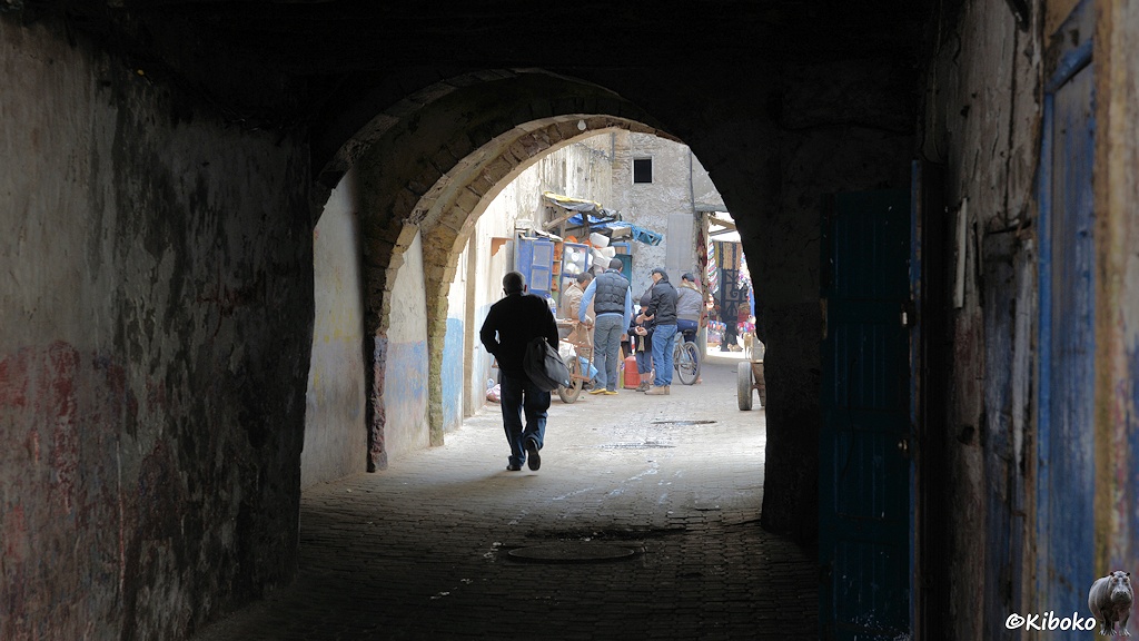 Das Bild zeigt einen Durchgang mit Rundbogen einer Gasse. Ein Mann läuft gerade durch den Bogen. Dahinter steht eine Gruppe Männer vor einem Geschäft, dessen Waren bis in die Gasse ragen.