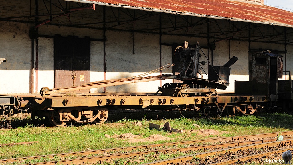 Das Bild zeigt einen vierachsigen gelblich verrosteten Flachwagen mit einem verrosteten grauen Bremserhaus. Darauf steht ein zweiachsiger Kranwagen mit herunterhängenden Ausleger.