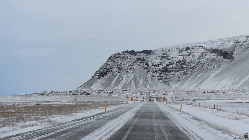 Das Bild zeigt eine Teestraße über die Seitwärts der Wind den Schnee weht. Die Straße führt gerade auf einen Berg zu. An der Straße sind gelb-rote Warnschilder auf beiden Seiten. In größerer Entfernung ist eine schmale Brücke mit nur einer Fahrspur.