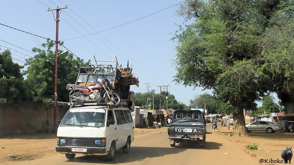 Das Bild zeigt einen weißen Kleinbus auf einer staubigen Teerstraße in einr Ortschaft. Links ist eine hohe Mauer. Rechts steht ein großer Baum. Voraus fährt ein Geländewagen. Der Kleinlaster ist hoch beladen. Vorn steht ein Motorrad auf dem Dach.
