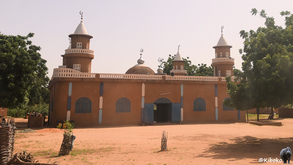Das Bild zeigt eine rotbraune Moschee mit hellem Geänder auf dem Dach. An den Ecken sind vier runde Türme. In der Mitte ist eine runde Kuppel. Die blaue Doppelflügeltür steht weit offen. Die vier vergitterten Fenster sind geschlossen.
