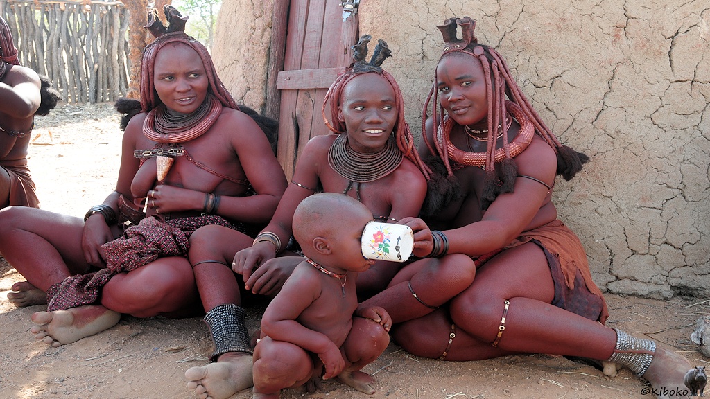 Drei Frauen sitzen vor einer Hütte im Schatten. Einem kleinen Jungen wird gerade zu trinken gegeben.