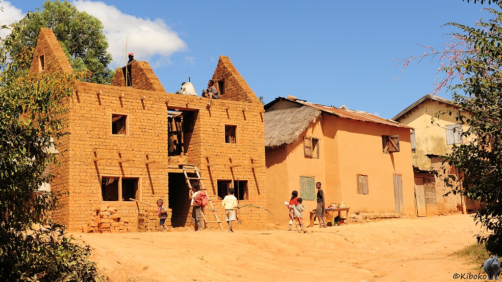 Das Bild zeigt ein zweistöckiges Haus im Rohbau. Die Wände bestehen aus rotbraunen Lehmziegeln. Eine einfache Holzleiter führt in den ersten Stock.