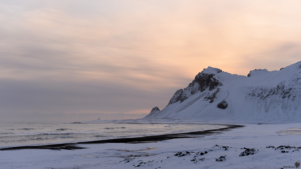 Das Bild zeigt eine kleine Bucht im Gegenlicht. Die Landschaft ist mit Schnee bedeckt. Ein schwarzer Strandstreifen ist an der Küste. In der rechten Bildhälfte verdeckt eine Bergkette die tiefstehende Sonne. Am Ende der Bergkette steht ein kleiner Leuchtturm am Wasser.