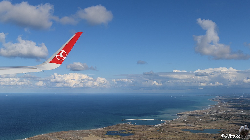 Das Bild zeigt eine Luftaufnahme auf eine Küste am blauen Meer. Eine Tragfläche mit einem roten Winglet ragt von links ins Bild.