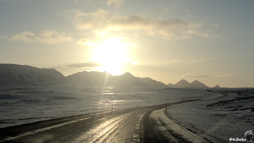 Die Sonne steht über einer verschneiten Bergkette. Eine vereiste Teerstraße führt an der Bergkette entlang