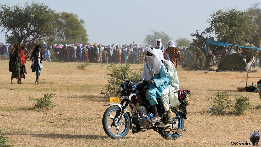 Das Bild zeigt zwei Männer mit türkisem und blassgrünem Gewand und weißen Turbanen auf einem Motorrad. Im Hintergrund laufen drei Frauen zu einer größeren Menschenansammlung.