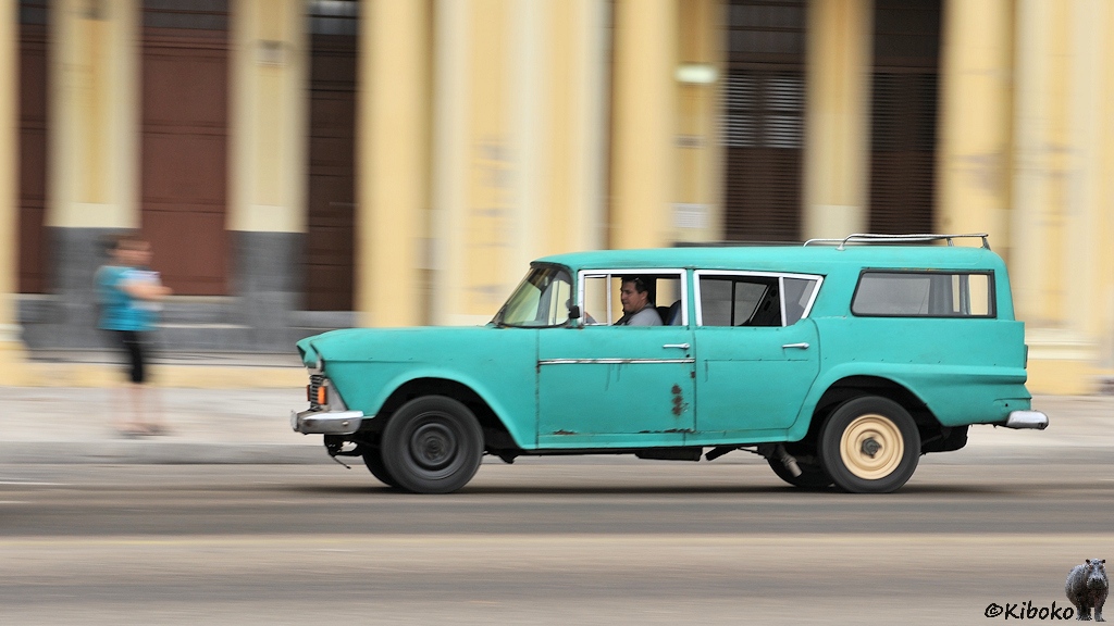 Das Bild zeigt einen Mitzieher eines türkisen alten amerikanisches Autos. Es ist ein Kombi, wobei der hintere Teil sich zwichen die Heckflossen quetscht. Die Fenster sind nicht auf einer Linie. Ein restliches Stück Chromleiste ziert die Fahrertür. Im Hintergrund ist ein unscharfes blassgelbes Haus.