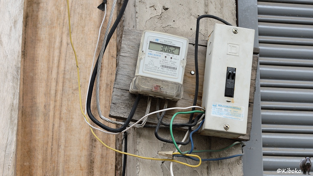 Das Bild zeigt einen digitalen Sromzähler. Links daneben ist eine hellgraue Kiste mit zwei Schaltern. Beides ist auf ein morsches Brett montiert, welches mit einem Nagel an eine Außenwand eines Hauses befestigt ist. Die elektrischen Leitungen baumeln an den Geräten.