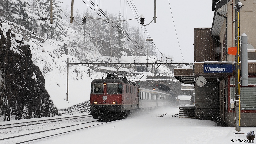 Das Bild zeigt eine rote Elektrolokomotive mit einem Personenzug aus grün-weißen Wagen bei der Ausfahrt aus einen Tunnel im Schneetreiben.