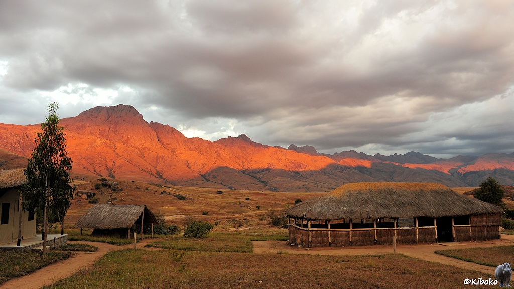 Das Bild zeigt eine große strohgedeckte, offene Hütte an einer vertrockneten Rasenfläche. Dahinter ist ein Tal. Die dahinterliegende Bergkette wird von der tiefstehenden Sonne rotorange angestrahlt. Darüber iehen graue Wolken.