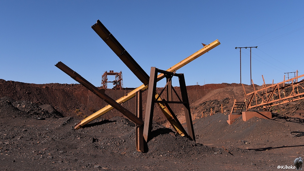 Ein rostiges, gelbes Gestänge steht im dunkelgrauen Schotter. Im Hintergrund sind weitere Reste der Verladeanlage zu sehen.