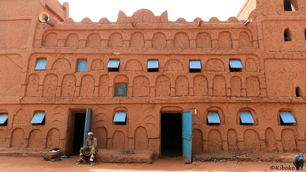 Das Bild zeigt den Mittelteil der Moschee mit zwei offenen, hellblauen Türen. Die Fenster sind durch hellblaue Fensterladen geschützt, die geöffnet unten von der Wand abstehen. Vorder linken Tür sitzt ein Mann im graugrünen Gewand. Links davon stehen vier paar Flip-Flops und ein paar Turnschuhe.