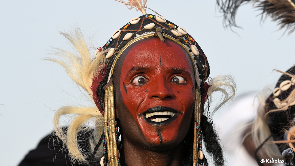 Das Bild zeigt das Porträt eines Mannes mit rötlich gefärbtem Gesicht, schwarzen Lippen und schwarzen auf der Stirn und am Kinn. Er zeigt die weißen Zähne und die weit aufgerissenen Augen schielen.